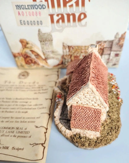A miniature model of a cottage in Inglewood from the Lilliput Lane collection, displayed with its instruction booklet and packaging box on a light blue background.