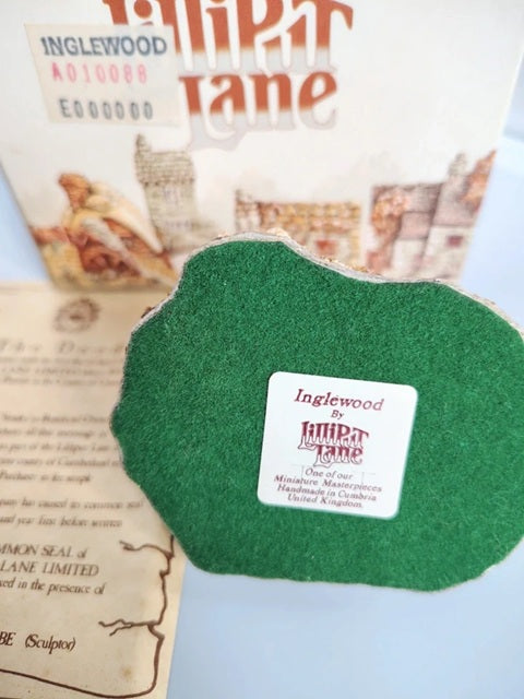 A green felt-shaped Lilliput Lane cottage miniature accessory with a white product label, displayed in front of its printed packaging box and instruction sheet on a light blue background. Inglewood Cottage.