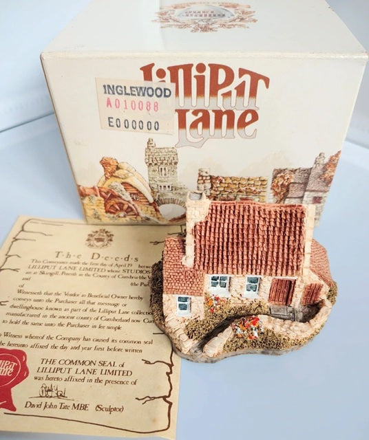 Inglewood Lilliput Lane cottage with its original box and instruction booklet on a light blue surface. The set includes a small brick-built house with a brown tiled roof, a miniature figure, and a detailed printed box showing the company branding and illustrations.