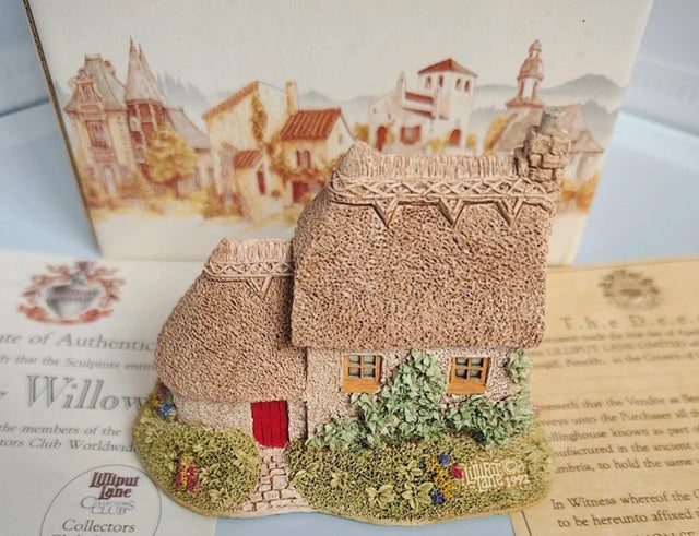 A miniature decorative cottage figurine with textured thatched roofs, green shrubs, and a small red door, displayed in front of printed cards featuring a village scene.