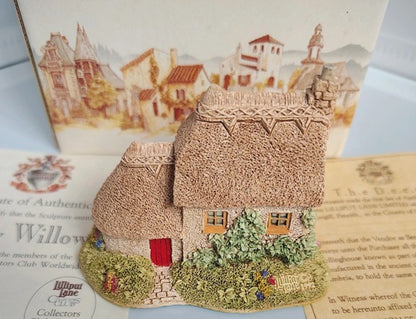 A miniature decorative cottage figurine with textured thatched roofs, green shrubs, and a small red door, displayed in front of printed cards featuring a village scene.