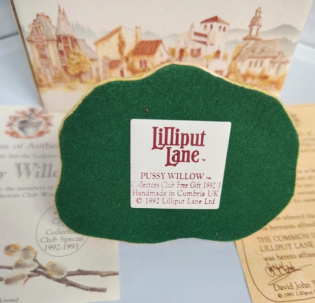 A green felt Lilliput Lane 'Pussy Willow' collector's club free gift, shown with its printed packaging and inserts in the background.