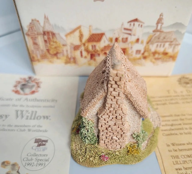 A miniature decorative cottage figurine with a textured beige stone facade and a green grassy base, displayed in front of its illustrated packaging box and authenticity certificate.