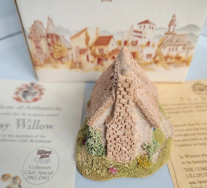 A miniature decorative cottage figurine with a textured beige stone facade and a green grassy base, displayed in front of its illustrated packaging box and authenticity certificate.