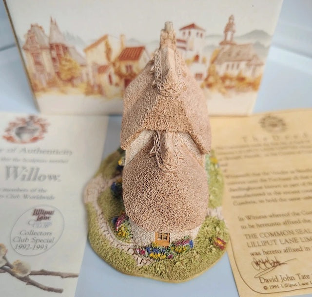 A miniature decorative cottage figurine with a textured beige roof and a small colorful garden base, displayed with printed authenticity cards and a scenic illustration card in the background.