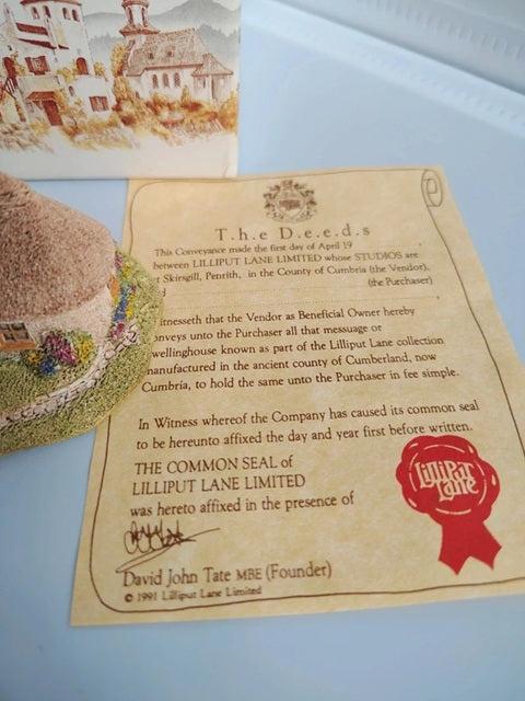 A vintage-style Lilliput Lane Limited deed certificate on aged beige paper, shown alongside a small decorative illustration and a textured object in the background.