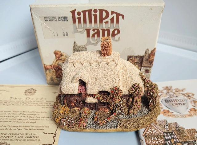 A miniature model of a cottage from the board game Lilipt Lane, displayed in front of its original box on a tabletop with printed deeds and booklet.
