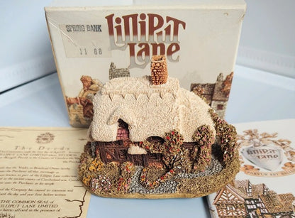A miniature model of a cottage from the board game Lilipt Lane, displayed in front of its original box on a tabletop with printed deeds and booklet.