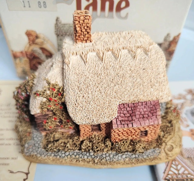 A miniature decorative Lilliput Lane Spring Bank cottage figurine with a textured cream thatched roof, brown chimney, and pinkish-brown tiled roof, displayed on a base with a blurred printed background.