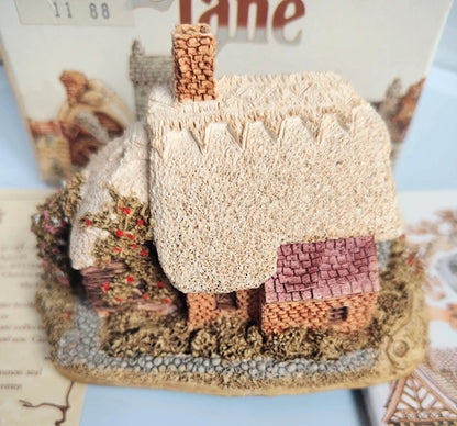 A miniature decorative Lilliput Lane Spring Bank cottage figurine with a textured cream thatched roof, brown chimney, and pinkish-brown tiled roof, displayed on a base with a blurred printed background.