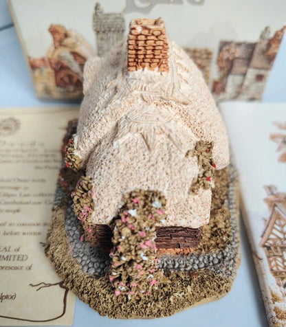 A miniature decorative gingerbread-style cottage model with a textured cream roof, brown chimney, and detailed base, displayed on a tabletop with vintage-style cards in the background.