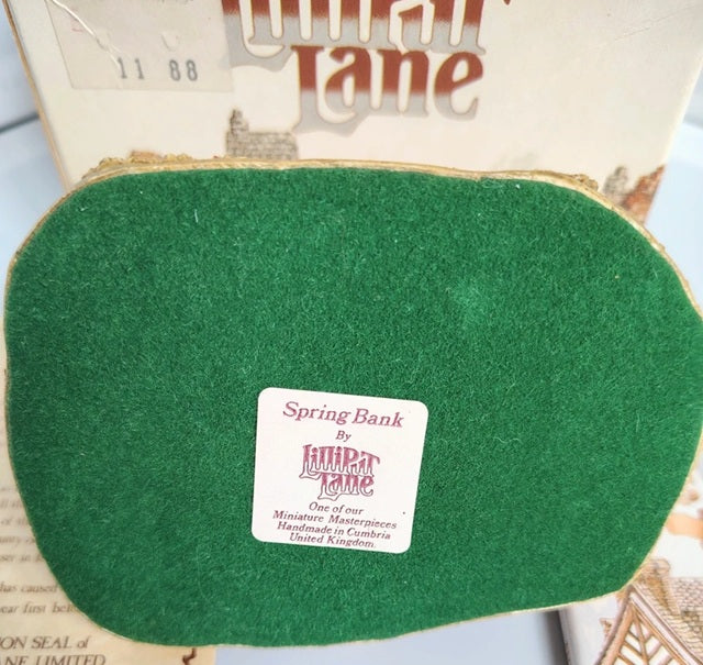 A vintage green felted wool Spring Bank by Lilliput Lane with a white product label in the center, set against a background of its original illustrated box.