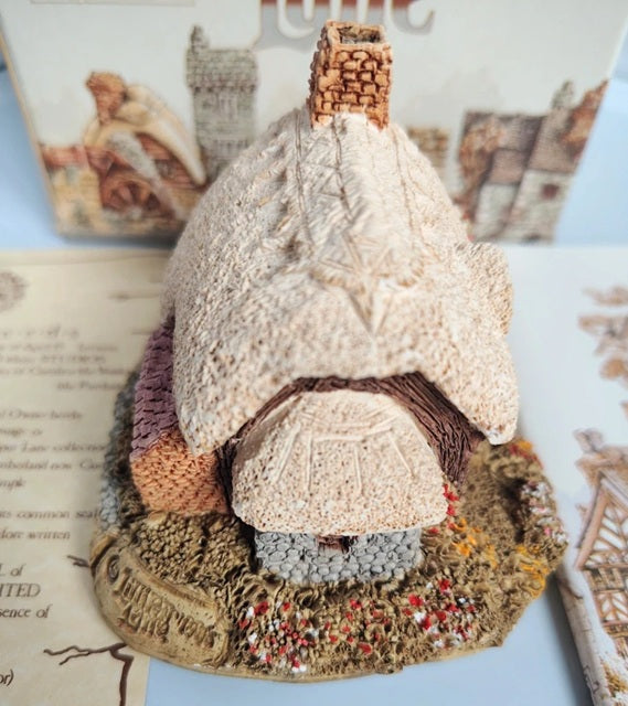A handcrafted crocheted cottage figurine with a textured cream-colored roof, brown chimney, and a multi-colored base, displayed on a light blue surface with vintage-style book pages in the background.