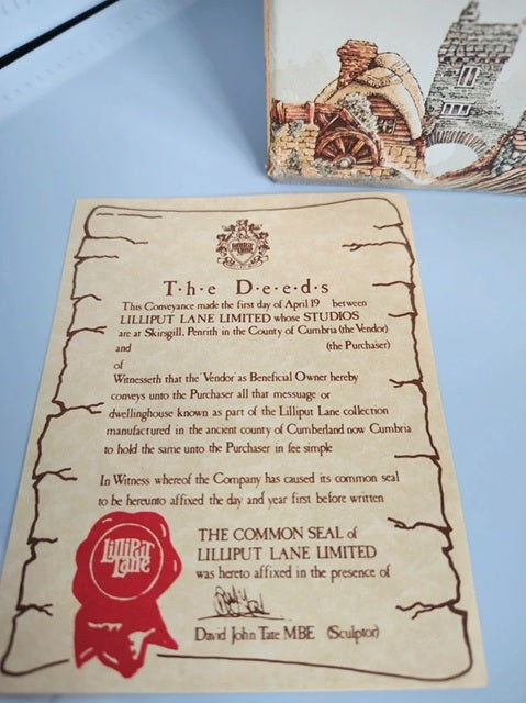 A replica of the fictional 'The Deeds' common seal from Lilliput Lane Limited, printed on aged parchment paper with a red wax-seal stamp, displayed on a light blue surface next to a matching illustrated card.