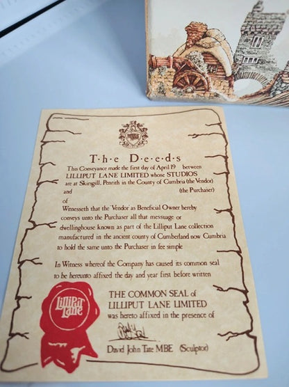 A replica of the fictional 'The Deeds' common seal from Lilliput Lane Limited, printed on aged parchment paper with a red wax-seal stamp, displayed on a light blue surface next to a matching illustrated card.
