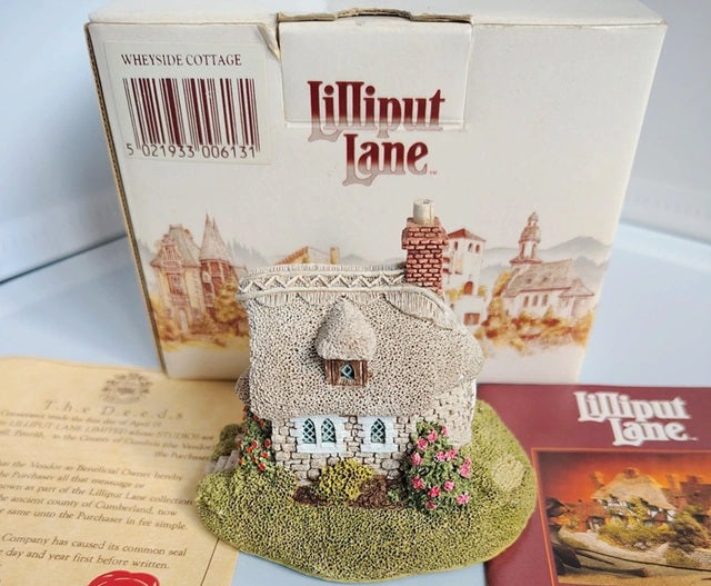 A miniature Wheyside Cottage from Lilliput Lane, displayed in front of its original white packaging box. The detailed model features a textured stone exterior, a small brown door, a green grassy base, and small pink flowers.