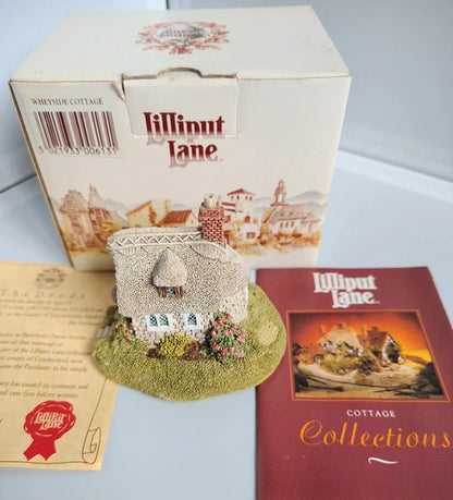 A Lilliput Lane 'Wheyside Cottage' ornament displayed with its original packaging box, an insert card, and a 'Lilliput Lane Cottage Collections' booklet on a light gray surface.