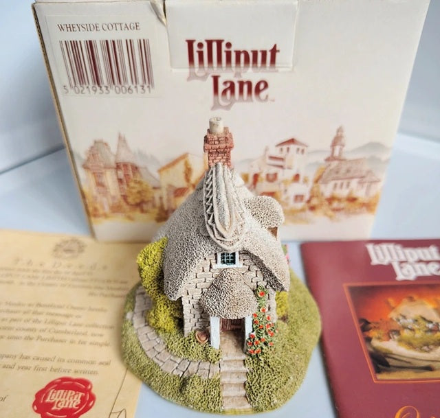 A miniature Lilliput Lane 'Wheyside Cottage' model displayed in front of its original packaging box, with a booklet and another card visible on a white surface.