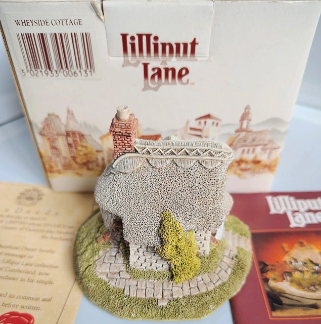 A miniature decorative cottage figurine from Lilliput Lane, displayed in front of its original white packaging box and next to a booklet, on a light blue surface.