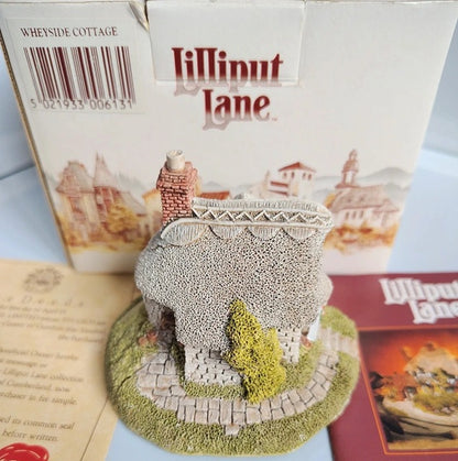 A miniature decorative cottage figurine from Lilliput Lane, displayed in front of its original white packaging box and next to a booklet, on a light blue surface.