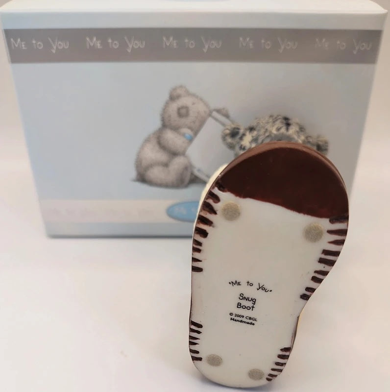 A Me to You 'Snug Boot' collectible figurine boot displayed in front of its original light blue packaging box. The boot features a white base with dark brown and black striped details, and a dark brown heel.
