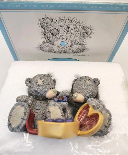 A Me To You teddy bear figurine set in its original white foam packaging, with the branded blue and white box lid visible behind it. The figurine depicts two grey textured teddy bears sitting together, one holding a small purple gift and the other opening a gold heart-shaped box filled with pink glittery contents.