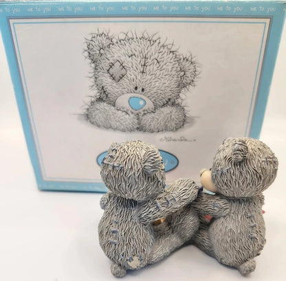 Two small textured teddy bear figurines hugging in front of a matching illustrated box on a white background.