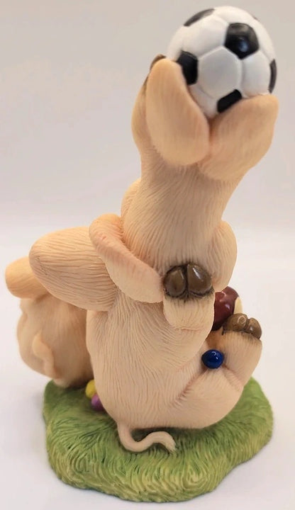 A whimsical figurine of a beige pig balancing on its back a black-and-white soccer ball, standing on a green grassy base. The figurine features detailed textures and small colorful accents, including a blue ball on the pig's back and tiny decorative elements.