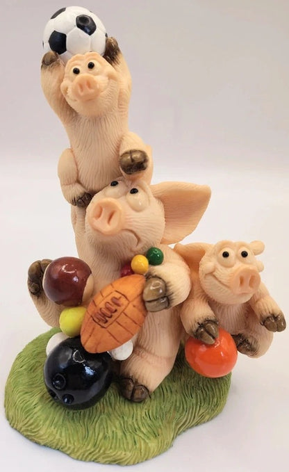 Piggin Balls Figurine by David Corbridge made in 1998.A whimsical figurine of three stacked pigs playing with various sports balls on a green grass-like base, set against a plain light background.