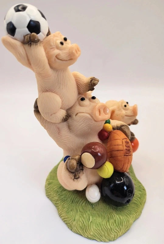 A whimsical figurine of three stacked pigs balancing various sports balls on their backs, set on a green grass-like base against a plain light background.