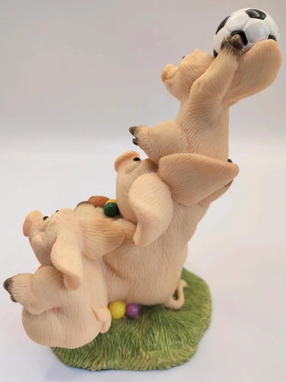 A whimsical figurine of three stacked pigs balancing a soccer ball overhead, set on a green grassy base with small colorful balls, photographed against a plain light background.