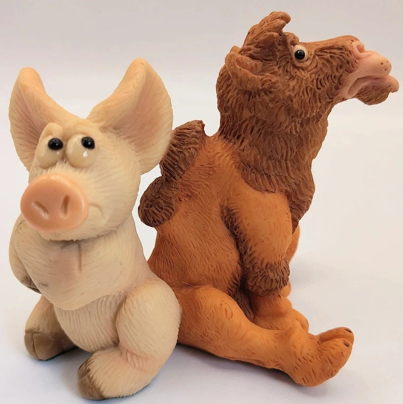 Piggin Got The Hump figurine by David Corbridge made in 2003. Two small sculpted figurines on a plain light background: a pale cream pig facing forward and a brown camel-like creature turned partly away, both with textured surfaces and black bead eyes.