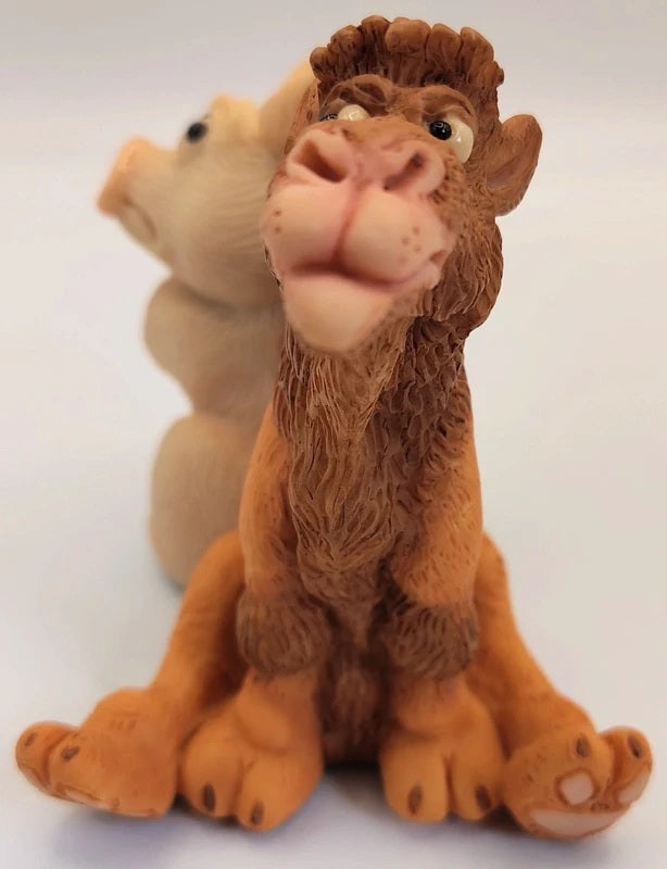 A small camel figurine in warm brown and tan tones, posed sitting against a plain light background, with a second pig animal figure blurred behind it.