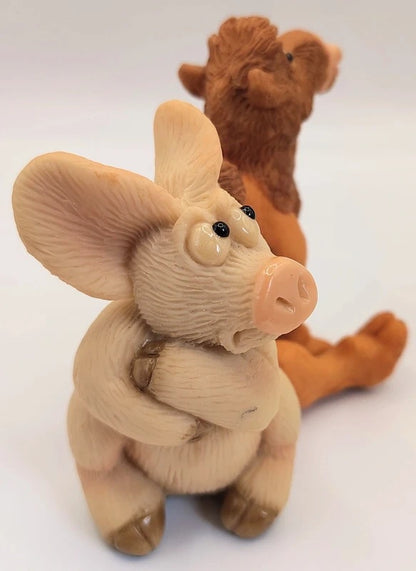 A small sculpted pig figurine in the foreground with a brown camel figurine behind it on a plain white studio background.