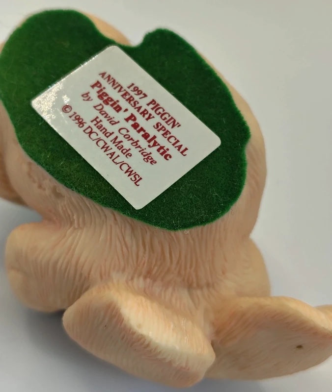 A close-up of a vintage 1997 DCC 'Piggin' Paralytic' collectible figurine, showing the back side with a white paper label attached to its green felt patch.