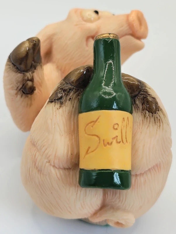 A small figurine of a pig holding a green bottle with a yellow label that reads 'Swill' in script, photographed against a plain white background.