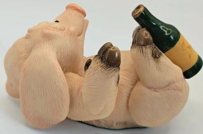 A novelty figurine of a pig lying on its back, holding a green and yellow wine bottle in its paws, photographed against a plain white background.