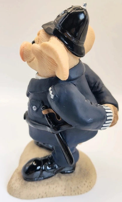 A small figurine of a pig dressed as a police officer, wearing a black helmet and dark blue uniform with a black baton, standing on a beige textured base against a plain white background.