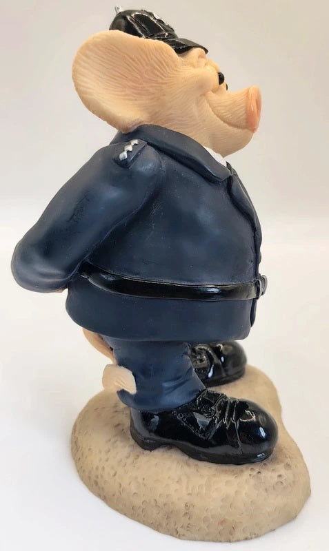 A collectible figurine of a pig dressed in a dark navy blue police uniform with a black hat, belt, and boots, standing on a textured beige base against a white background.