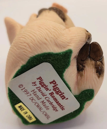 A vintage 1997 collectible figurine of a pig's feet, featuring textured beige skin, dark brown hooves, and a green felt patch with a white label reading 'Piggin' Romantic by David Corbridge'.