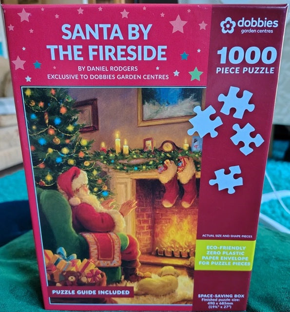 Box for a 1000‑piece jigsaw puzzle titled “Santa By The Fireside,” showing Santa sitting in a green armchair beside a decorated fireplace and Christmas tree, with festive stockings, candles and garlands in a warm holiday scene.