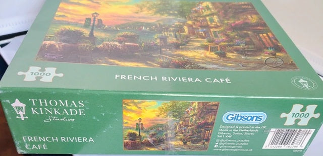 Jigsaw puzzle box featuring a scenic design with 'French Riviera Café' text and Thomas Kinkade branding.