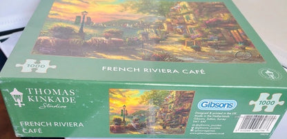 Jigsaw puzzle box featuring a scenic design with 'French Riviera Café' text and Thomas Kinkade branding.