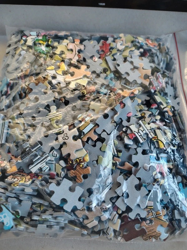 A clear plastic bag filled with many small printed jigsaw puzzle pieces, photographed from above on a light surface.