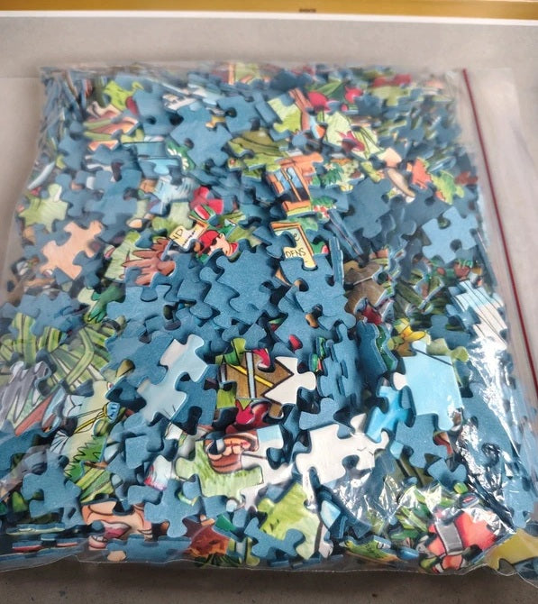 A clear plastic bag filled with many small jigsaw puzzle pieces featuring a colorful cartoon-style landscape scene, photographed from above on a light surface.