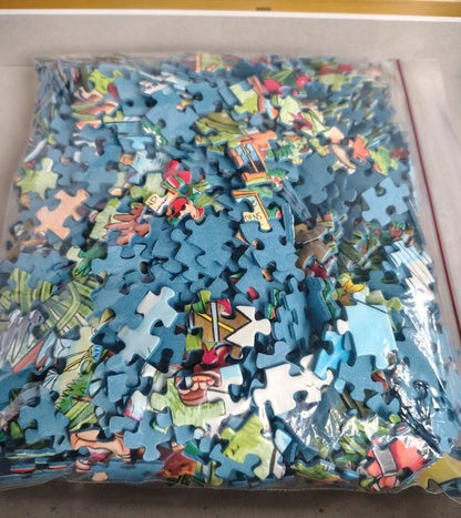 A clear plastic bag filled with many small jigsaw puzzle pieces featuring a colorful cartoon-style landscape scene, photographed from above on a light surface.